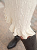 Close-up of a cream cable-knit skirt with a ruffled hem, paired with black leather boots, shown outdoors on a stone pavement.