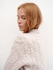 A woman with shoulder-length strawberry blonde hair is shown from the back, wearing a chunky cream-colored cable-knit sweater against a textured white wall.