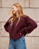 A woman with long blonde hair wearing a fuzzy, oversized deep burgundy sweater and blue jeans, standing against a light beige stone wall.