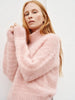 A woman with long blonde hair wearing a fuzzy, oversized light pink knit sweater, posing against a textured white wall.