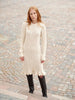 A woman with red hair wearing a cream cable-knit long-sleeve midi dress with a ruffled hem, paired with black knee-high leather boots, standing on a cobblestone street.