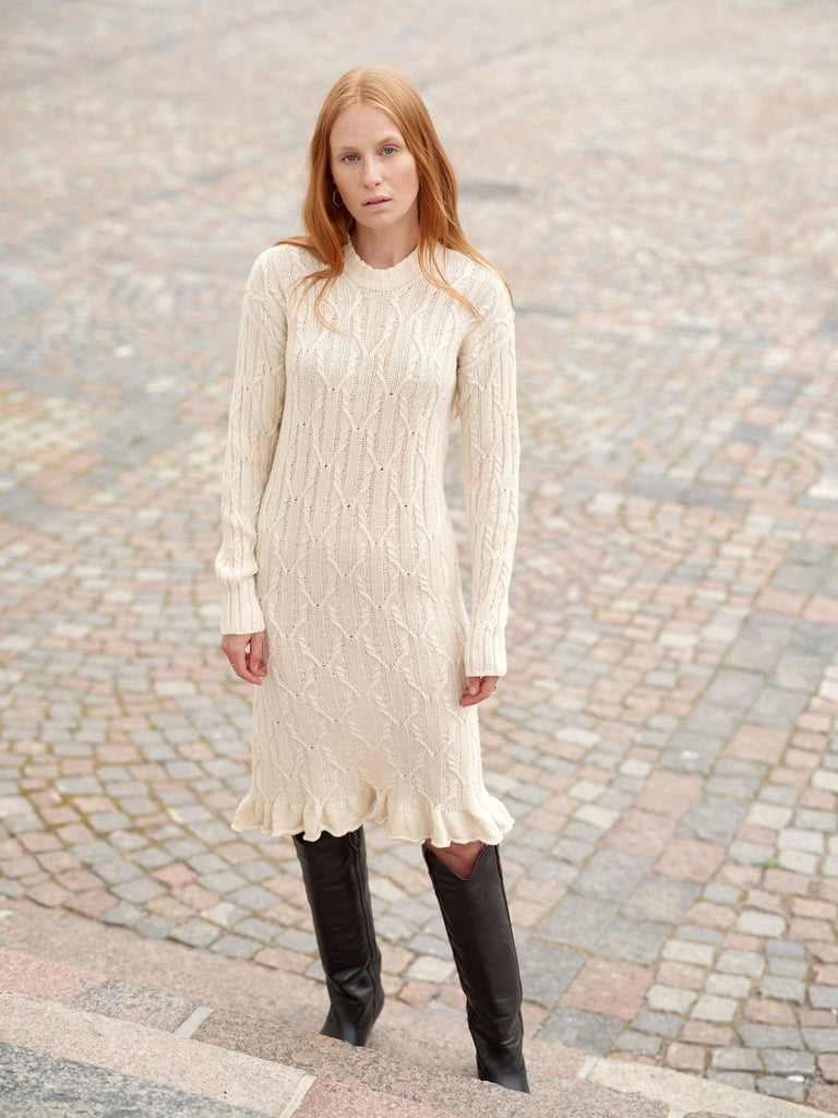 A woman with red hair wearing a cream cable-knit long-sleeve midi dress with a ruffled hem, paired with black knee-high leather boots, standing on a cobblestone street.