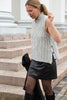 A woman wearing a light gray cable-knit sleeveless sweater vest, a black faux-leather mini skirt, sheer black tights, and black boots, standing on stone steps.