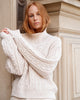A woman with strawberry-blonde hair wearing a chunky cream cable-knit mock-neck sweater, posing in front of a beige stone building.