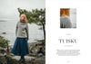 Woman wearing a patterned sweater and blue skirt standing by a lake with trees in the background.