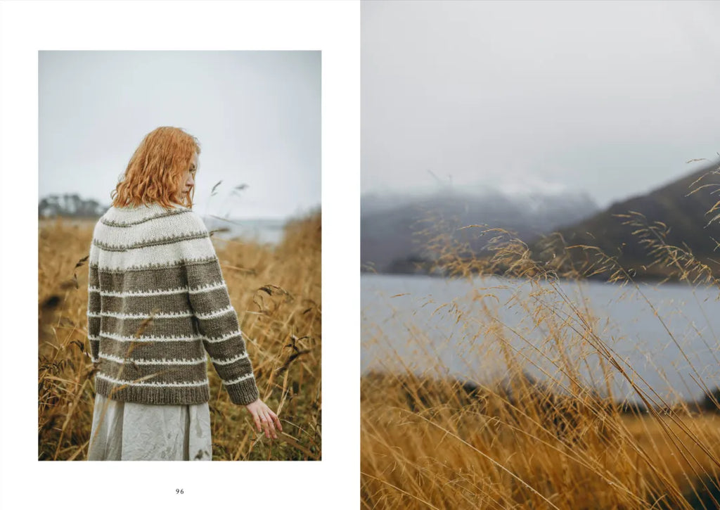 Person in a striped sweater standing in a field with a lake and mountains in the background