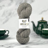 A skein of gray yarn with a white paper label reading 'KNIT WITH ORIGINS' stands upright on a light marble surface, with dark green teacups and a teapot blurred in the background.