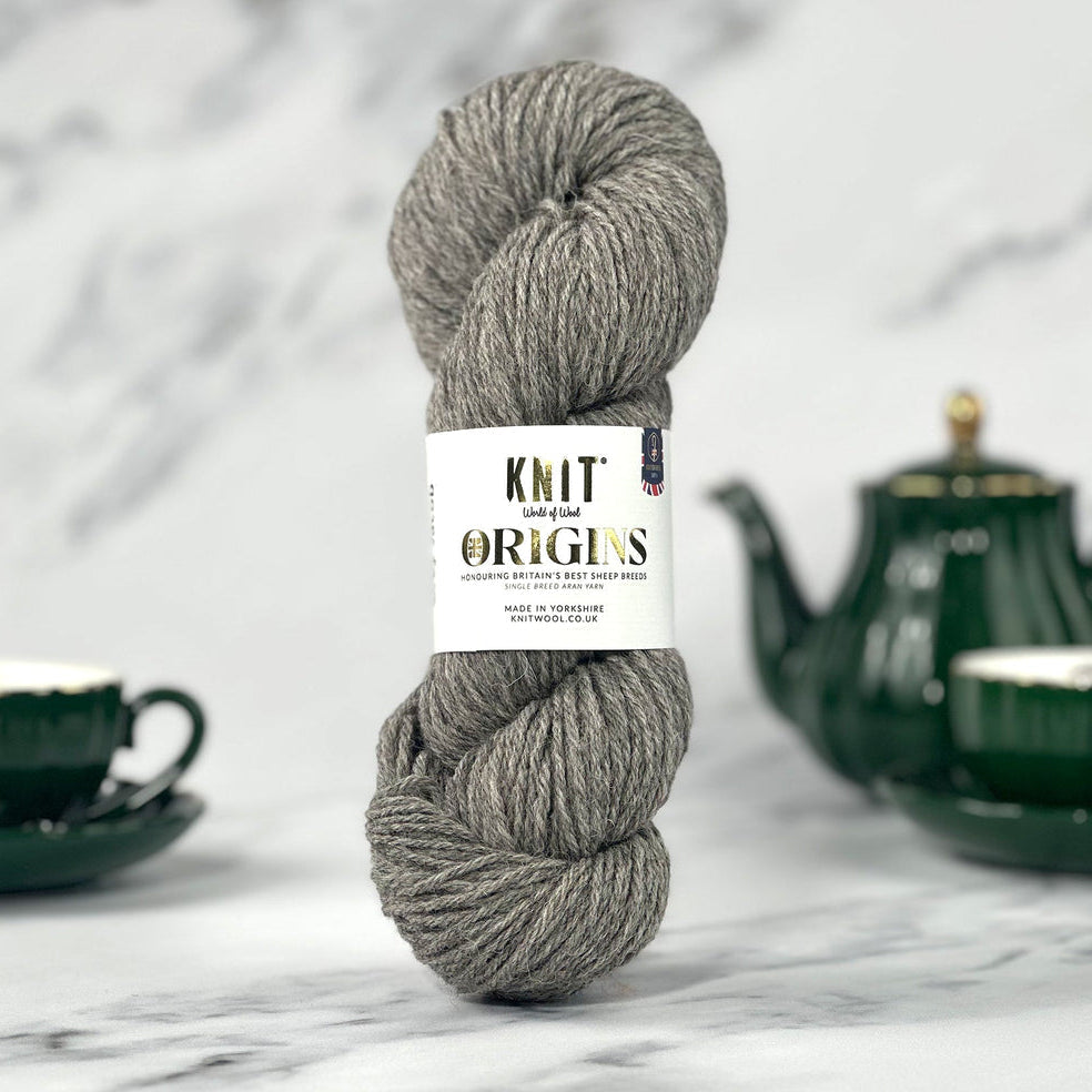 A skein of gray yarn with a white paper label reading 'KNIT WITH ORIGINS' stands upright on a light marble surface, with dark green teacups and a teapot blurred in the background.