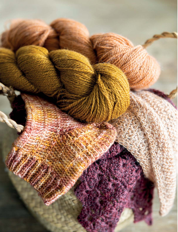 A close-up of variegated yarn skeins in warm autumn shades, including mustard yellow, peach, cream, and plum, draped over knitted swatches of matching yarn.