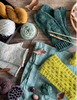 A styled flat-lay of knitted textile items on a rustic wooden surface, including a pair of dark green knit mittens, a folded cream knit piece, a bright yellow knit scarf, several balls of yarn, wooden knitting needles, dried leaves, and acorns.