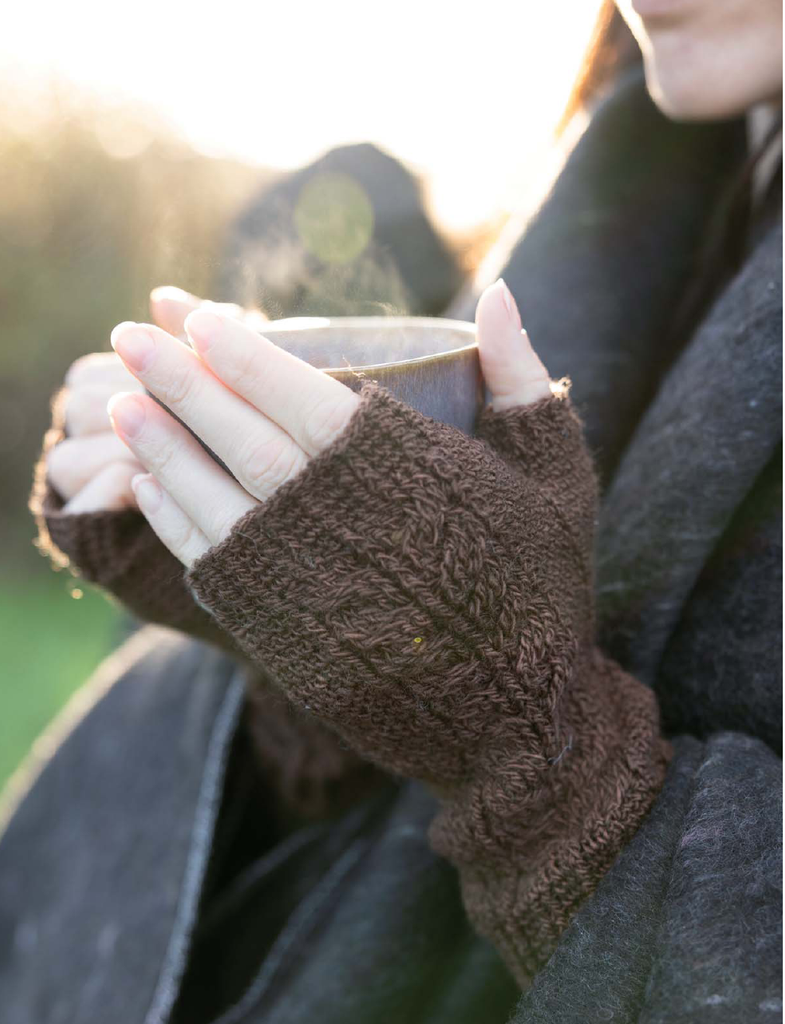 A close-up lifestyle shot of a person wearing a dark brown chunky knit fingerless glove while holding a steaming mug outdoors in warm sunlight.