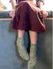 A person sitting on a bench wearing chunky knit ankle socks in a heathered olive green and cream yarn with a rust-colored dress.