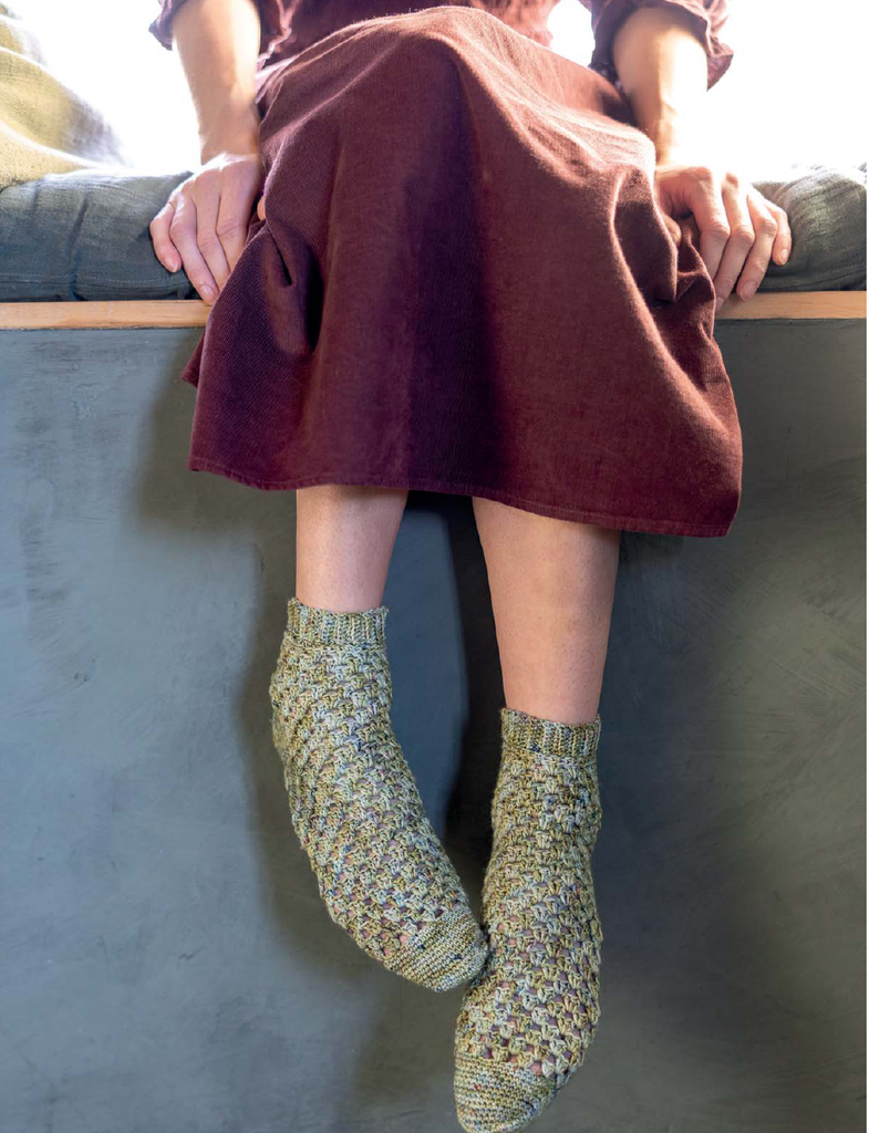 A person sitting on a bench wearing chunky knit ankle socks in a heathered olive green and cream yarn with a rust-colored dress.