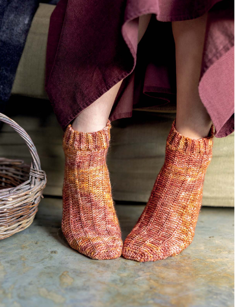 A person wearing a pair of hand-knit ankle socks in a variegated orange, yellow, and pink yarn, with a dusty mauve skirt and a wicker basket visible in a cozy indoor setting.