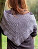A woman viewed from behind wearing a gray triangular knitted shawl draped over her shoulders, with a dark brown top underneath and an outdoor blurred background.