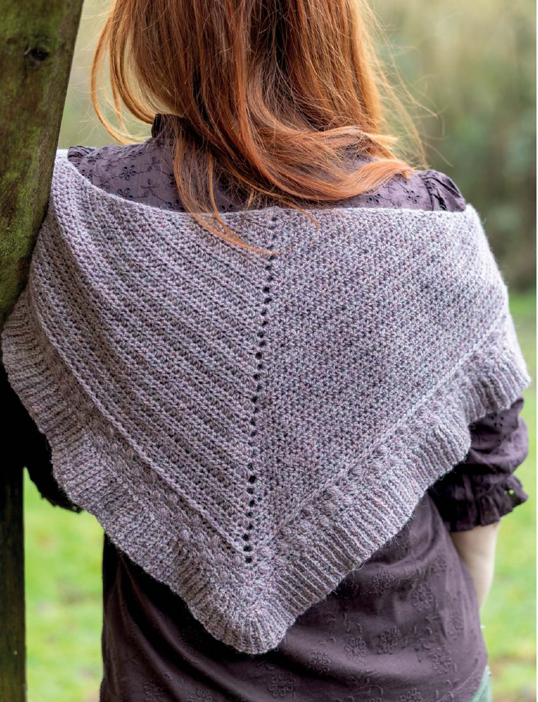A woman viewed from behind wearing a gray triangular knitted shawl draped over her shoulders, with a dark brown top underneath and an outdoor blurred background.