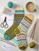Two knitted pattern socks displayed on wooden hangers with knitting needles, yarn balls, scissors, a cup of coffee, and pattern sheets arranged on a light marble surface.