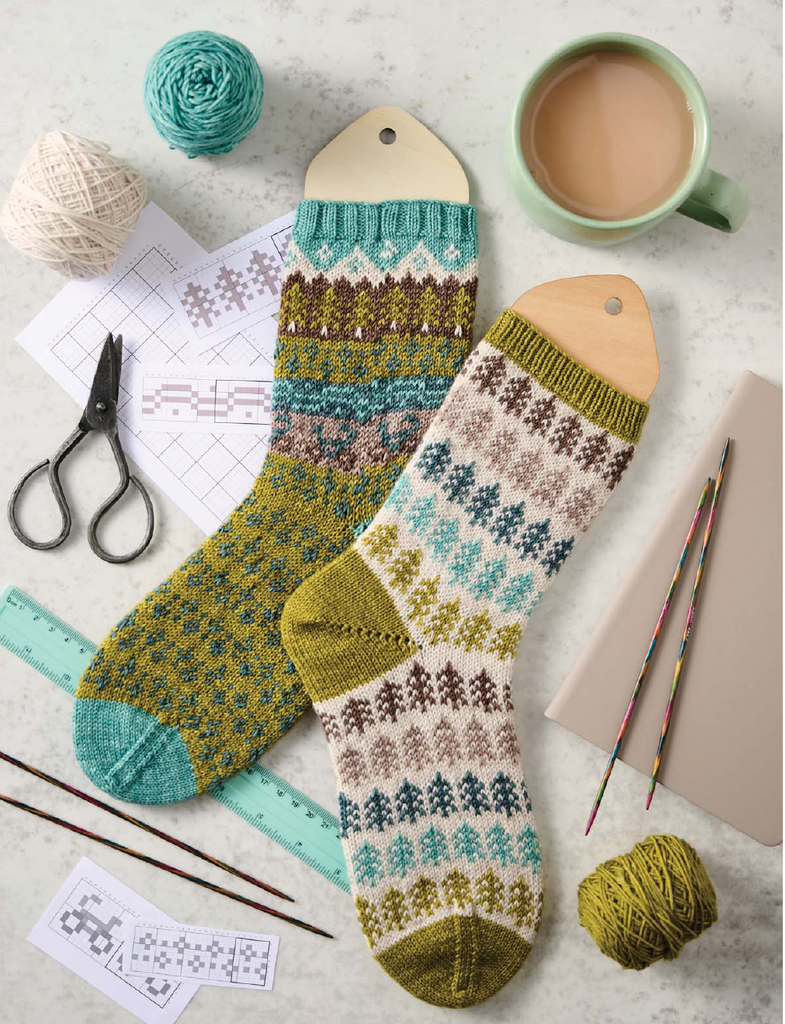 Two knitted pattern socks displayed on wooden hangers with knitting needles, yarn balls, scissors, a cup of coffee, and pattern sheets arranged on a light marble surface.