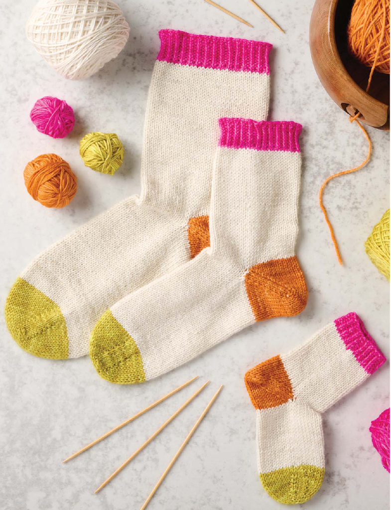 Hand-knit socks with cream bodies, bright pink ribbed cuffs, orange heels, and yellow toes, styled with knitting needles, yarn balls, and a wooden bowl on a light marble surface.