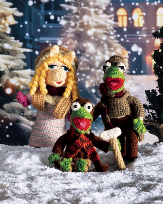 Three crocheted Muppet-style characters posed in a snowy outdoor scene with Christmas trees and a softly blurred building in the background. The front character holds a small axe, and all three have expressive faces and colorful yarn textures.