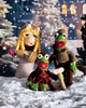 Three crocheted Muppet-style characters posed in a snowy outdoor scene with Christmas trees and a softly blurred building in the background. The front character holds a small axe, and all three have expressive faces and colorful yarn textures.