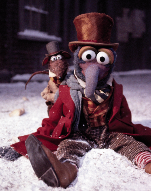 A plush puppet character with a long gray beak, large white eyes, and a brown top hat sits in the snow wearing a red coat and striped shirt, with a smaller matching puppet behind it. The scene is outdoors in snow with a dark building in the background.