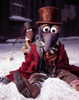 A plush puppet character with a long gray beak, large white eyes, and a brown top hat sits in the snow wearing a red coat and striped shirt, with a smaller matching puppet behind it. The scene is outdoors in snow with a dark building in the background.