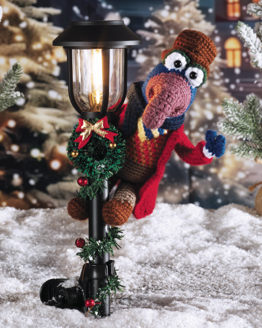 A crocheted holiday scene featuring a Muppet character in a red coat and brown hat leaning against a black solar-powered outdoor lamp decorated with a green wreath and red ornaments, set in a snowy festive background.