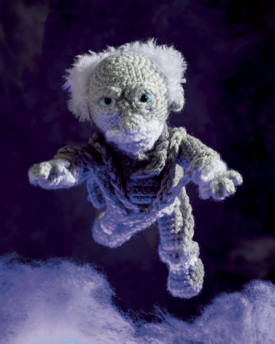 A crocheted amigurumi doll of a ghostly wizard or elf with white yarn hair, blue eyes, and a gray-and-white striped sweater, floating against a dark purple background with soft white cloud-like shapes at the bottom.