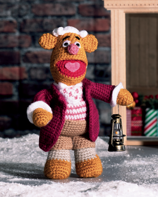 A crocheted Muppet-style character doll stands in a snowy scene, wearing a burgundy coat, white-and-red patterned sweater, tan pants, and holding a small lantern. A brick wall and wooden door are in the background.