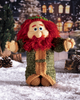A crocheted gnome doll with a red beard, beige face and hands, and an olive green and tan outfit, posed in a snowy winter village scene with pinecones and a warm-lit house in the background.
