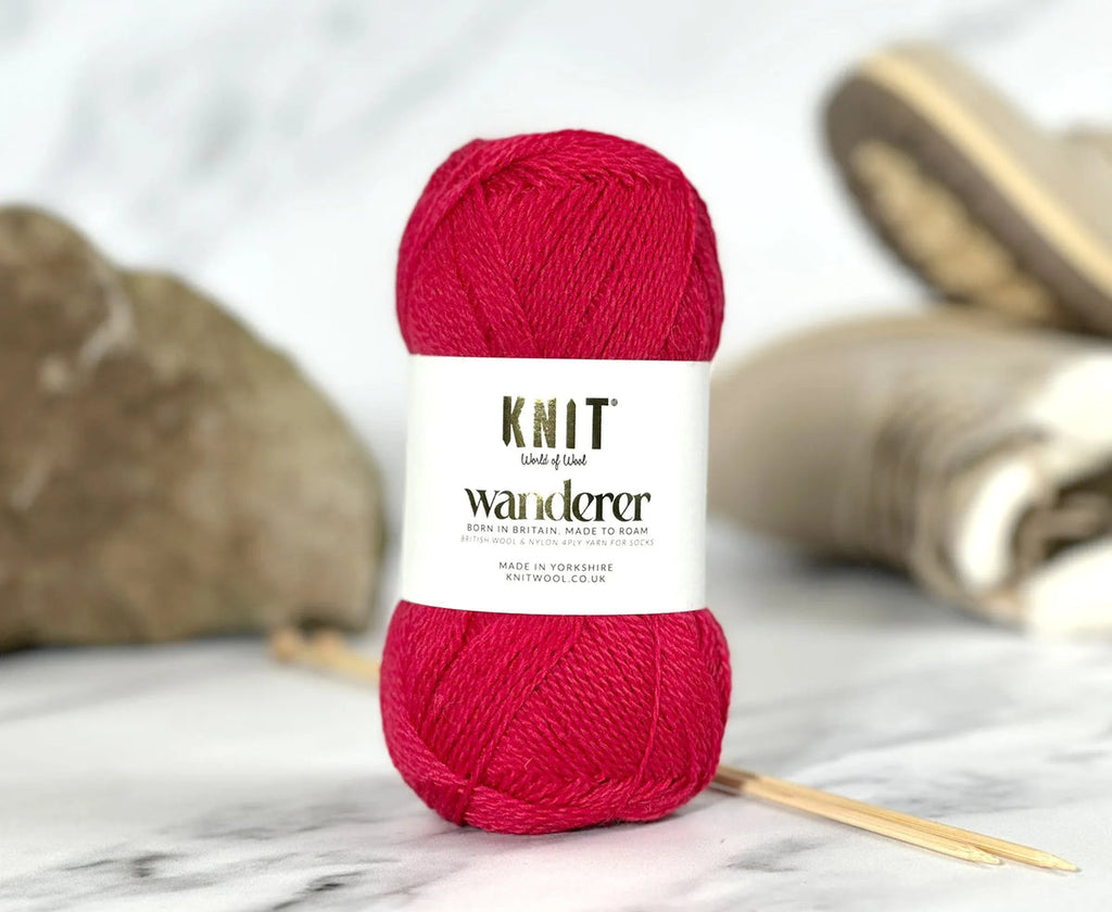 A skein of bright pink yarn with a white label reading 'KNIT lives of knit wanderer' is centered on a marble surface with knitting needles and soft blurred background props.