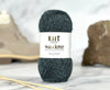 A skein of dark heathered gray yarn with a white label reading 'KNIT Wanderer' and 'Made in Britain Made to Measure,' photographed on a light marble-like surface with blurred beige and brown objects in the background.