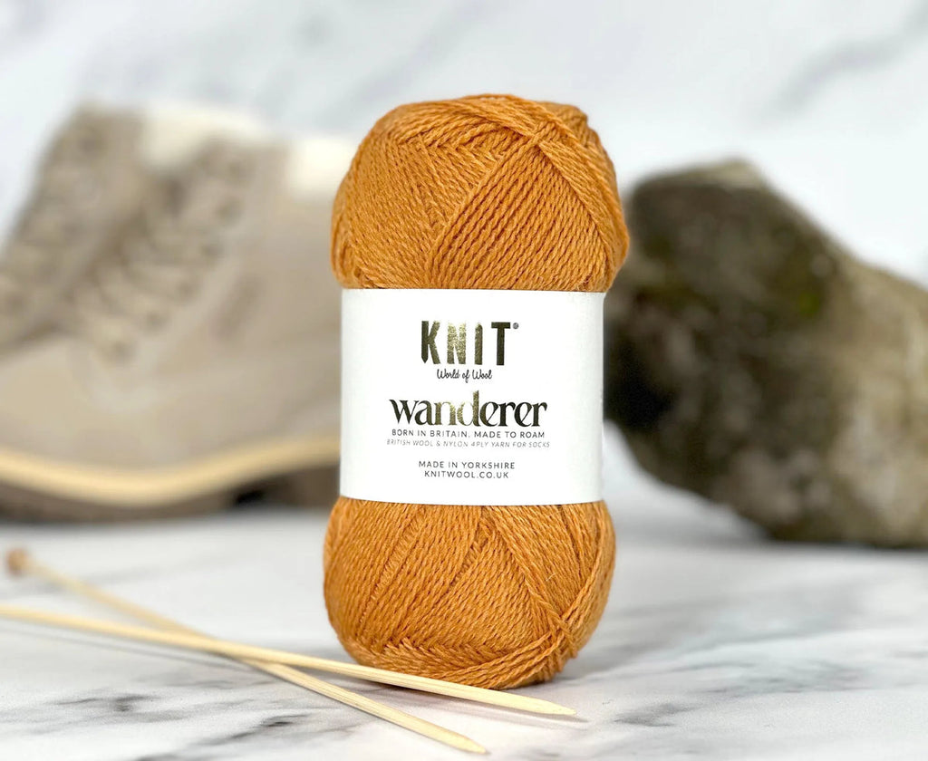 Ball of orange yarn with 'Knit Wanderer' label on a marble surface with knitting needles.