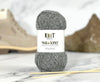 A ball of gray heathered yarn with a white label reading 'KNIT world of knit wanderer' is centered on a marble surface, with knitting needles in front and a blurred beige and brown background.