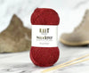 A ball of deep burgundy red yarn with a white paper label reading 'KNIT Wanderer' and 'Wanderer Yarn', photographed on a light marble surface with a blurred beige shoe and wooden knitting needles in the background.