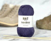 Purple yarn ball with 'Knit Wanderer' label on a marble surface with knitting needles.