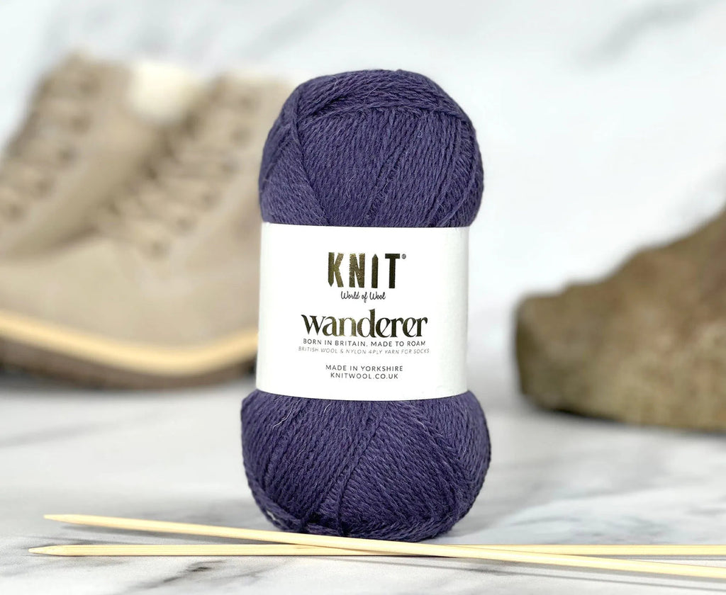 Purple yarn ball with 'Knit Wanderer' label on a marble surface with knitting needles.
