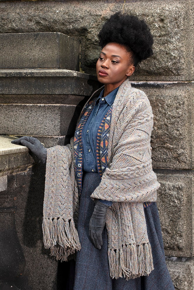 Woman wearing a patterned scarf and gloves leaning against a stone wall.