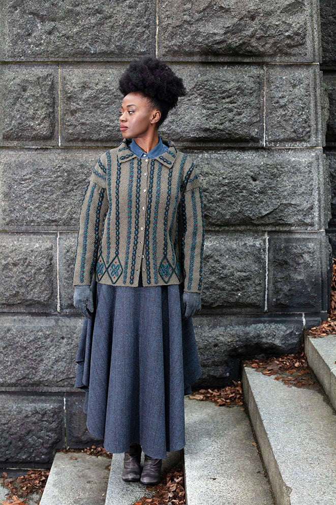 Woman wearing a patterned coat and long skirt standing against a stone wall.