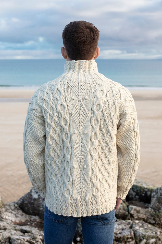 Man wearing a cream cable knit sweater standing on a beach with ocean view
