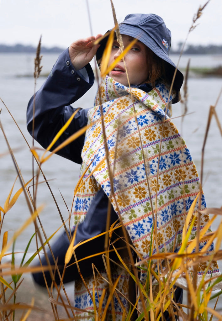Person wearing a patterned poncho and blue hat standing in tall grass by a body of water.