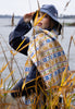 Person wearing a patterned poncho and blue hat standing in tall grass by a body of water.