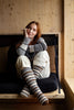 Woman sitting on a wooden bench wearing a gray sweater, white pants, and striped socks.