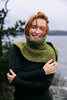 Woman wearing a green knitted cowl in a natural setting with water and trees.
