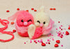 Two knitted characters, one pink and one white, holding a heart surrounded by small red and pink hearts on a light background.