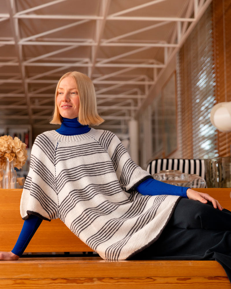Woman sitting on a wooden bench wearing a striped sweater and blue turtleneck in a modern indoor setting.
