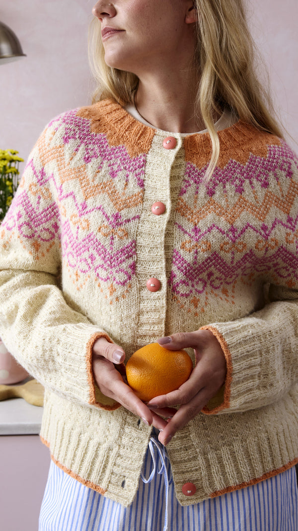 Person wearing a patterned cardigan holding an orange in a kitchen setting