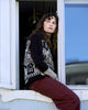 Woman wearing a patterned sweater and maroon pants sitting by a window.