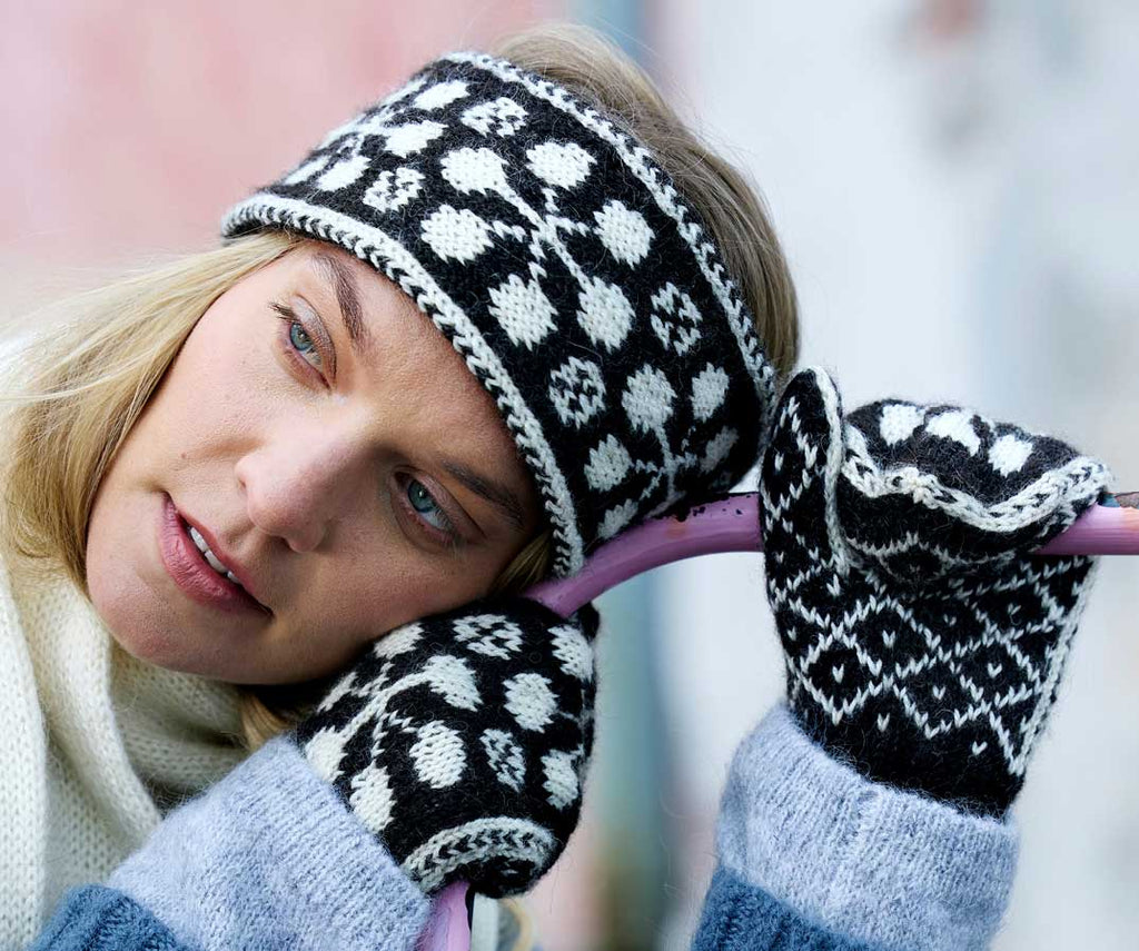 Person wearing a black and white patterned knitted hat and mittens, with a blurred background.